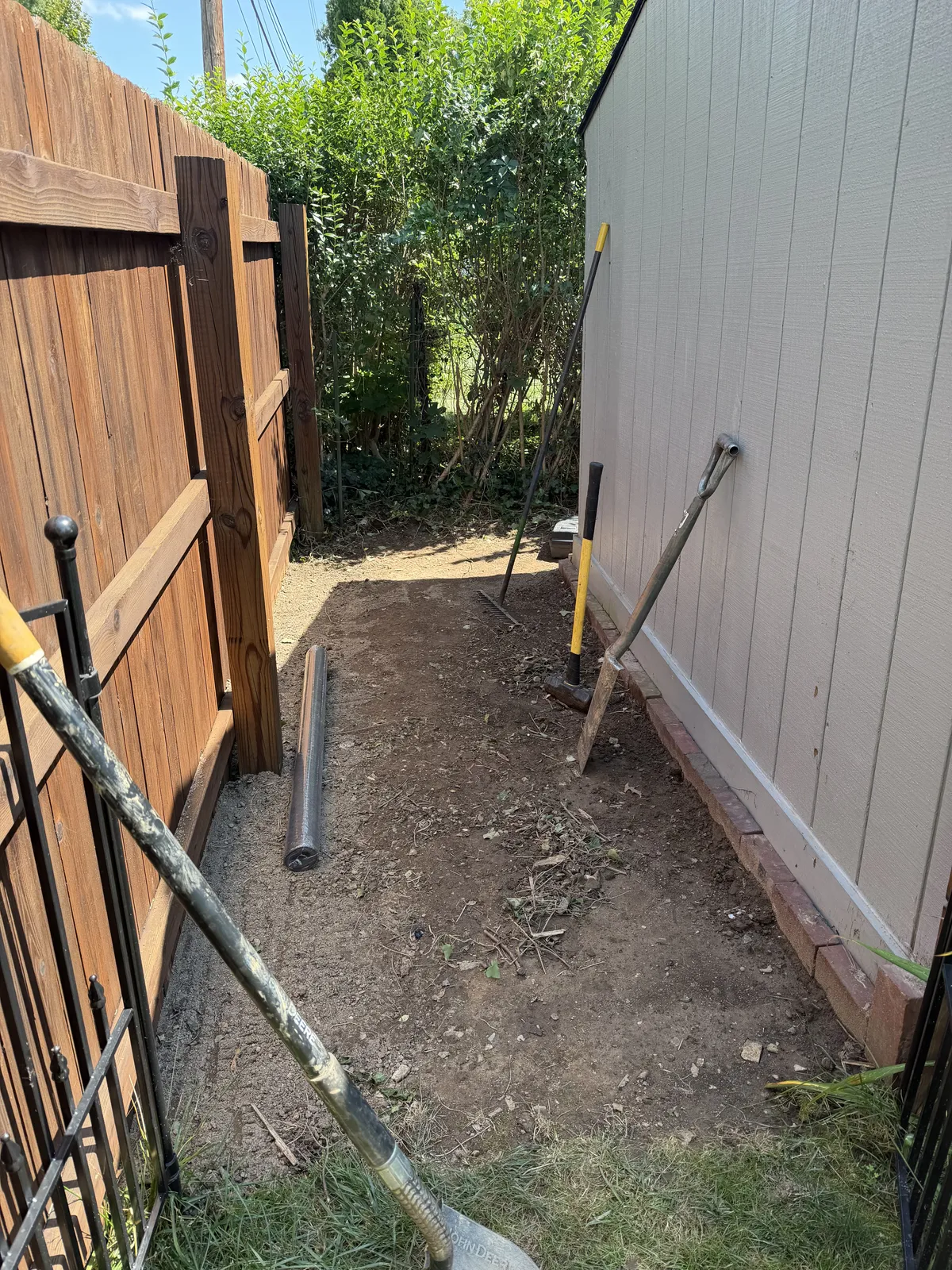 Cleared soil between a wooden fence and shed, with shovels, a rake, and digging tools positioned for ground preparation.