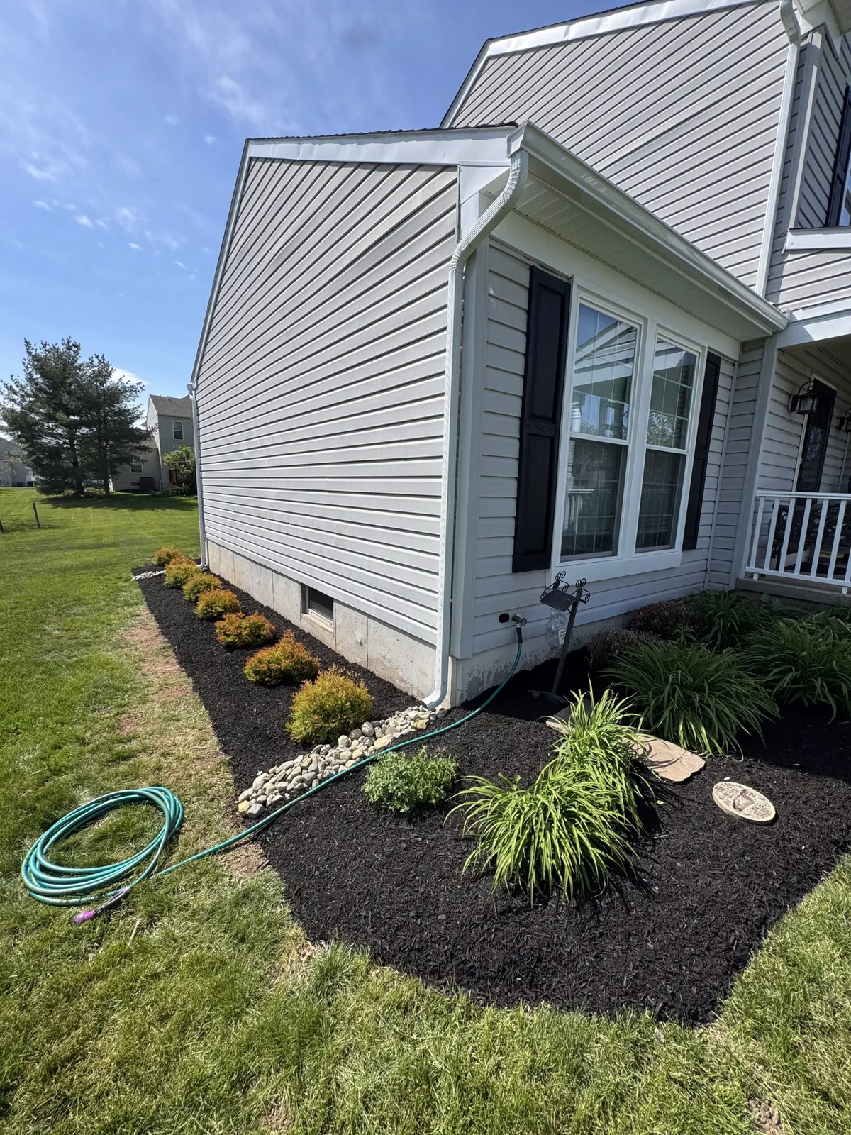 Curved mulch beds with neatly spaced shrubs, leafy plants, and a river rock strip beside a home’s foundation.