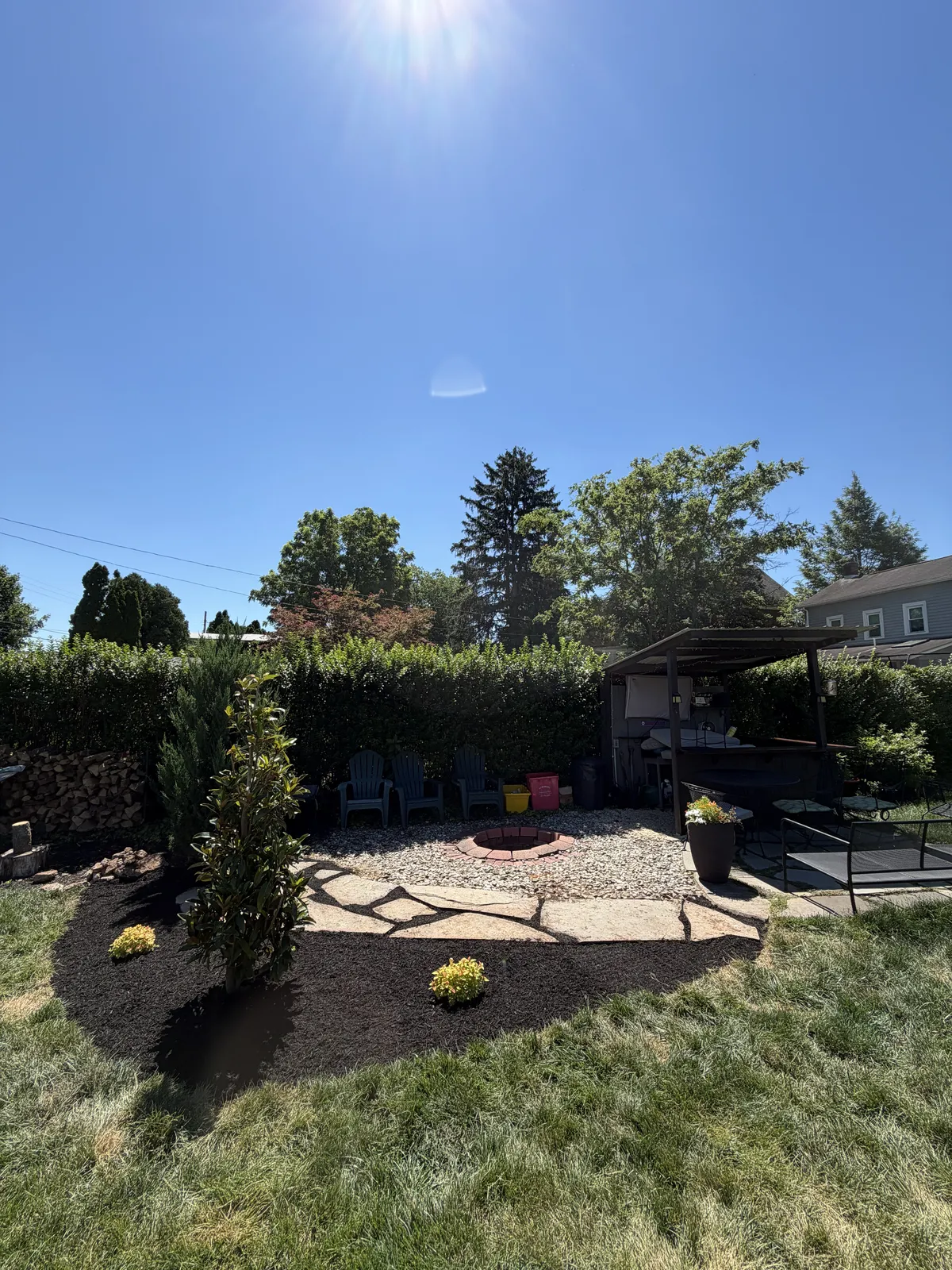 Backyard landscaping with a gravel fire pit area, flagstone border, flowering mulch beds, and privacy hedges.