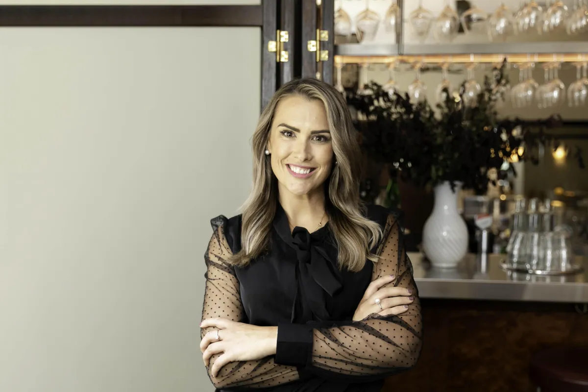 Portrait of Sarah Pagoda standing in a bright salon, smiling, with a bowl and brush on the counter in front of her.