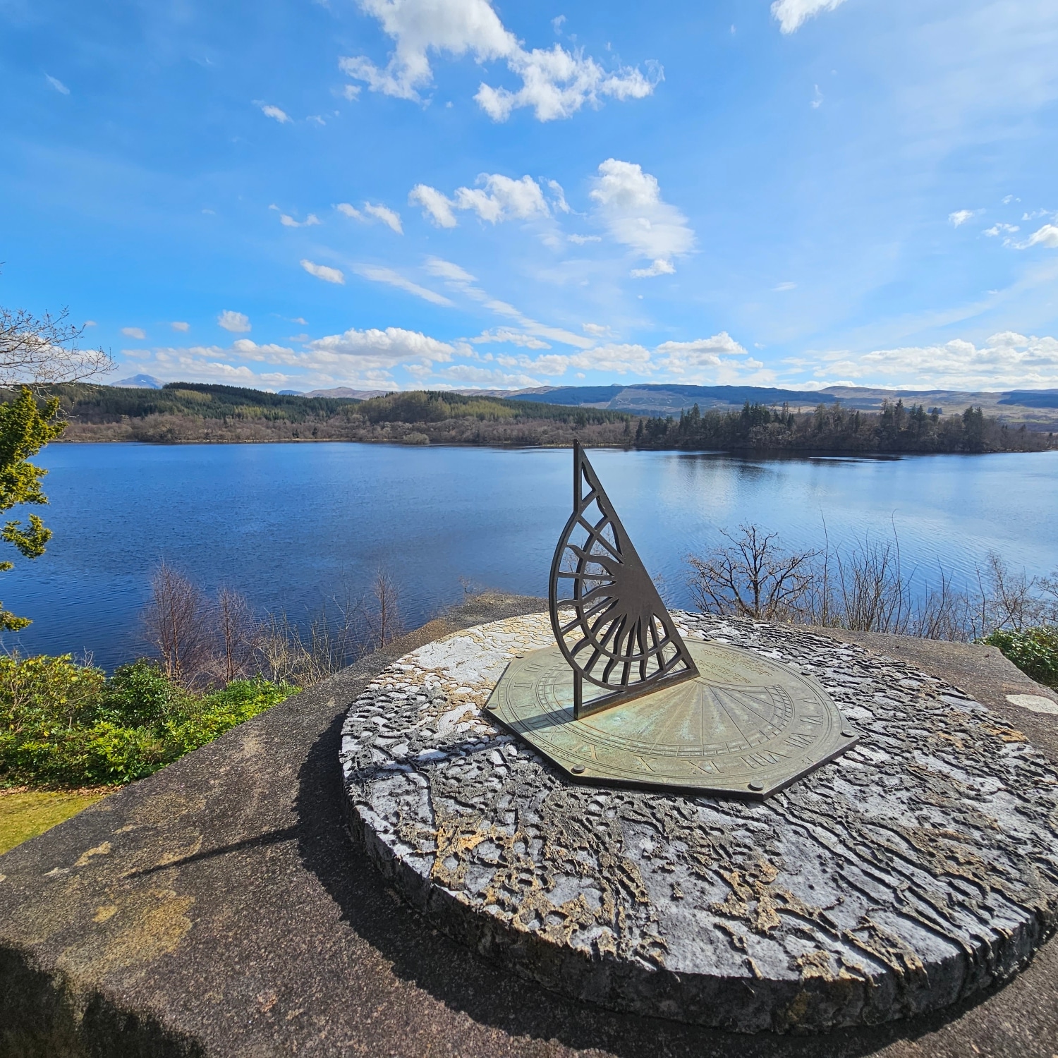 Stone sundial overlooking a calm blue lake and forested hills under a bright sky — real photograph taken in Scotland.