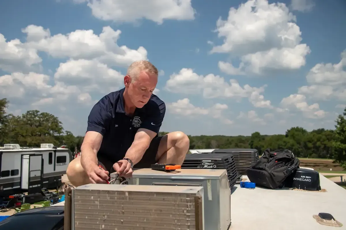 Technician performing RV air conditioning maintenance on rooftop, with tools and equipment visible, under a blue sky with clouds.
