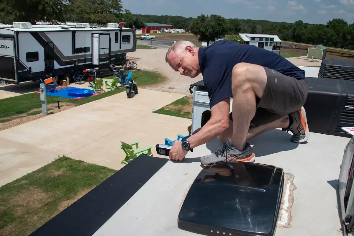 Man performing RV roof maintenance, inspecting and sealing roof components for water intrusion protection at a campsite.