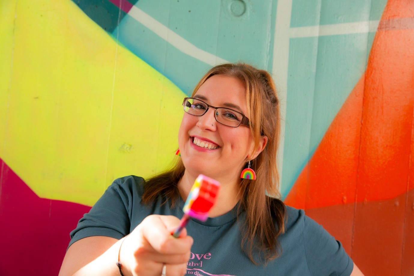 Smiling photo of Rachel holding a colourful pencil in front of a vibrant mural, representing creativity, experimentation and finding personalised strategies that support executive functioning and neurodivergent brains.