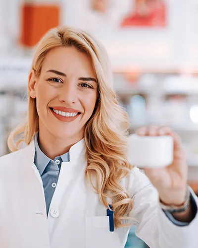 Apothekerin hält eine kleine Cremedose in die Kamera und lächelt, im Hintergrund Regale mit Produkten.