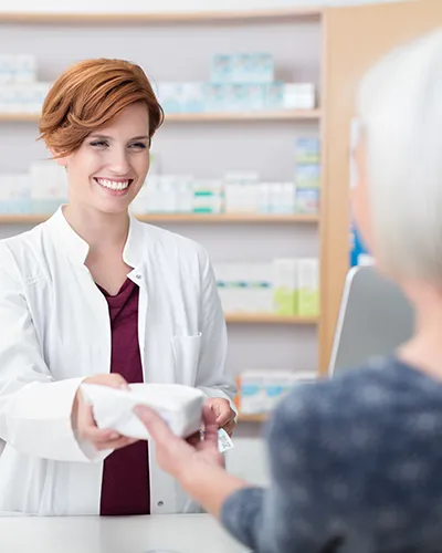 Apothekenmitarbeiterin reicht einer Kundin über die Kassentheke ein verpacktes Produkt und lächelt dabei freundlich