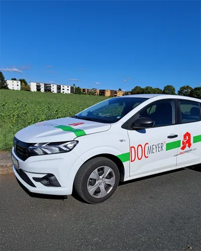 Ein weißes DOC Meyer Botendienst Auto mit grüner Streifengrafik und dem DOC Meyer Logo der Apotheke. Das Auto steht auf einer Straße, im Hintergrund sind Wohnhäuser und Bäume.
