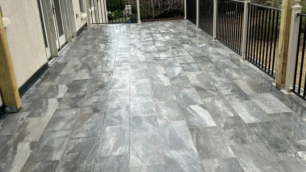 Spacious outdoor deck with gray stone tile flooring and black railing overlooking a wooded yard.