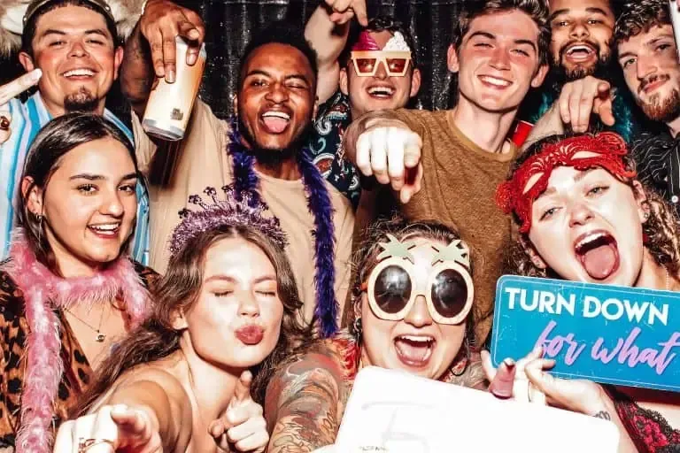 A group of people at a party, wearing colorful and playful accessories, smiling, pointing towards the camera, and holding a "Turn Down for What" sign. They're clearly enjoying the Photo Booth Rental DC experience!