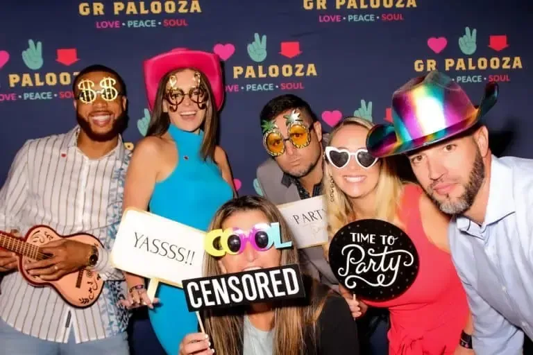 A group of six people pose playfully in front of a "GR Palooza" backdrop, wearing novelty glasses and hats, and holding prop signs with phrases like "Yasss!!", "Cool", and "Time to Party". This fun scene perfectly captures the excitement brought by a Photo Booth Rental.