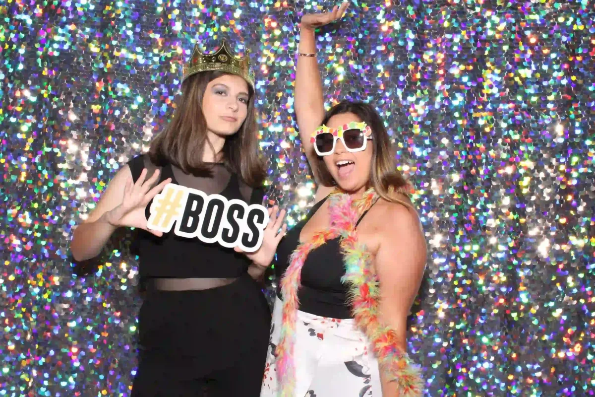 Two women pose playfully in front of a glittery backdrop at a party photo booth rental. One wears a crown and holds a "#BOSS" sign, while the other sports sunglasses and a feather boa, striking a celebratory pose.