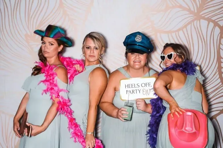 Four women in light green dresses pose for a photo booth rental. Two wear feather boas, one has a rainbow hat, another has a police hat, the third holds a sign saying "Heels Off, Party On," and the last one sports heart-shaped sunglasses and a pink cowboy hat.