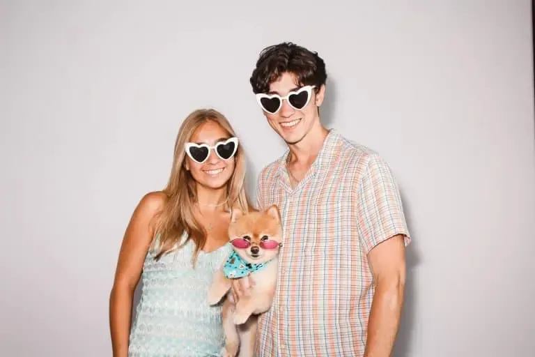 Two people stand smiling, each wearing heart-shaped sunglasses, while holding a small dog also sporting heart-shaped sunglasses and a bandana. The backdrop is plain white, perfect for a photo booth rental - capturing fun and memorable moments effortlessly.