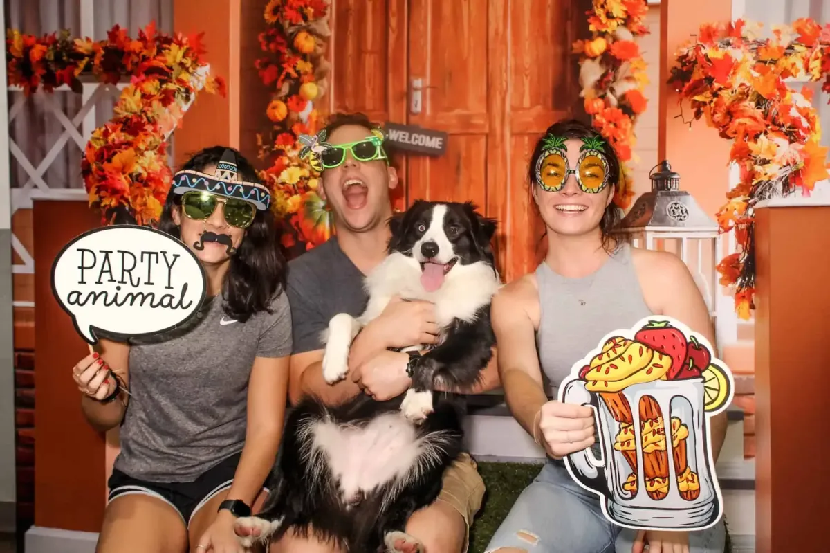 Three people in festive costumes pose with a dog in this lively private event. Two hold fun signs reading "Party Animal" with a beer mug and hot dog. An autumn-themed backdrop of orange leaves completes this photo booth snapshot.