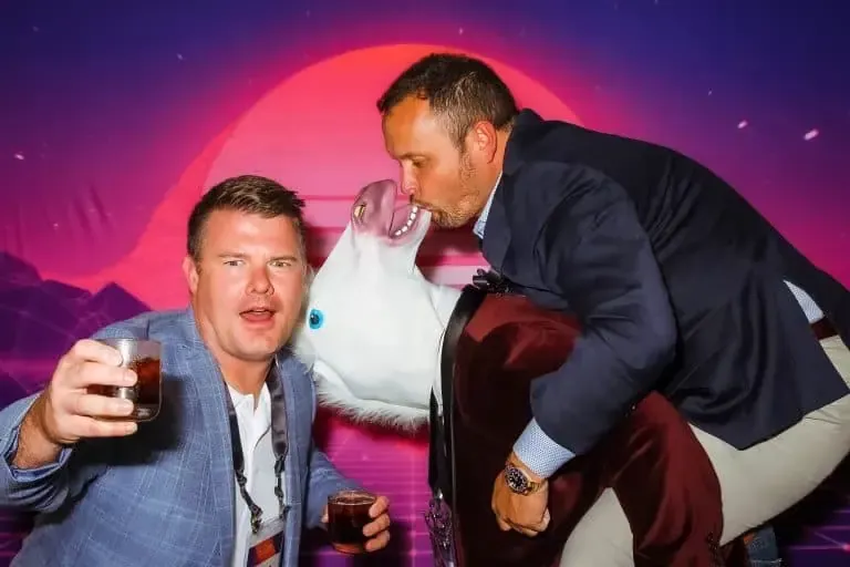 wo men in suits pose playfully in a Photo Booth; one holds a drink and looks at the camera, while the other kisses a person wearing a unicorn costume against a backdrop of a red sunset over mountains. Perfect for those considering Photo Booth Rental DC.