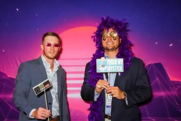 Two men in suits pose with retro-themed props and sunglasses at a corporate event. One holds a "TOUCH MY JOYSTICK" sign, the other an NES controller sign. Behind them, a pink and purple digital sunset sets the scene.