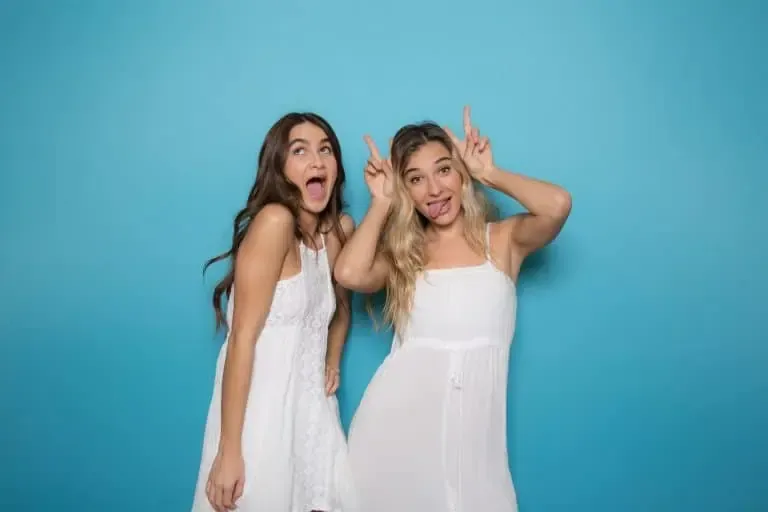 Two women in white dresses are posing playfully in front of a blue background, perfect for a fun photo booth session. One is making a goofy face while the other sticks out her tongue and makes bunny ears with her fingers. Ideal for any Photo Booth Rental DC event!