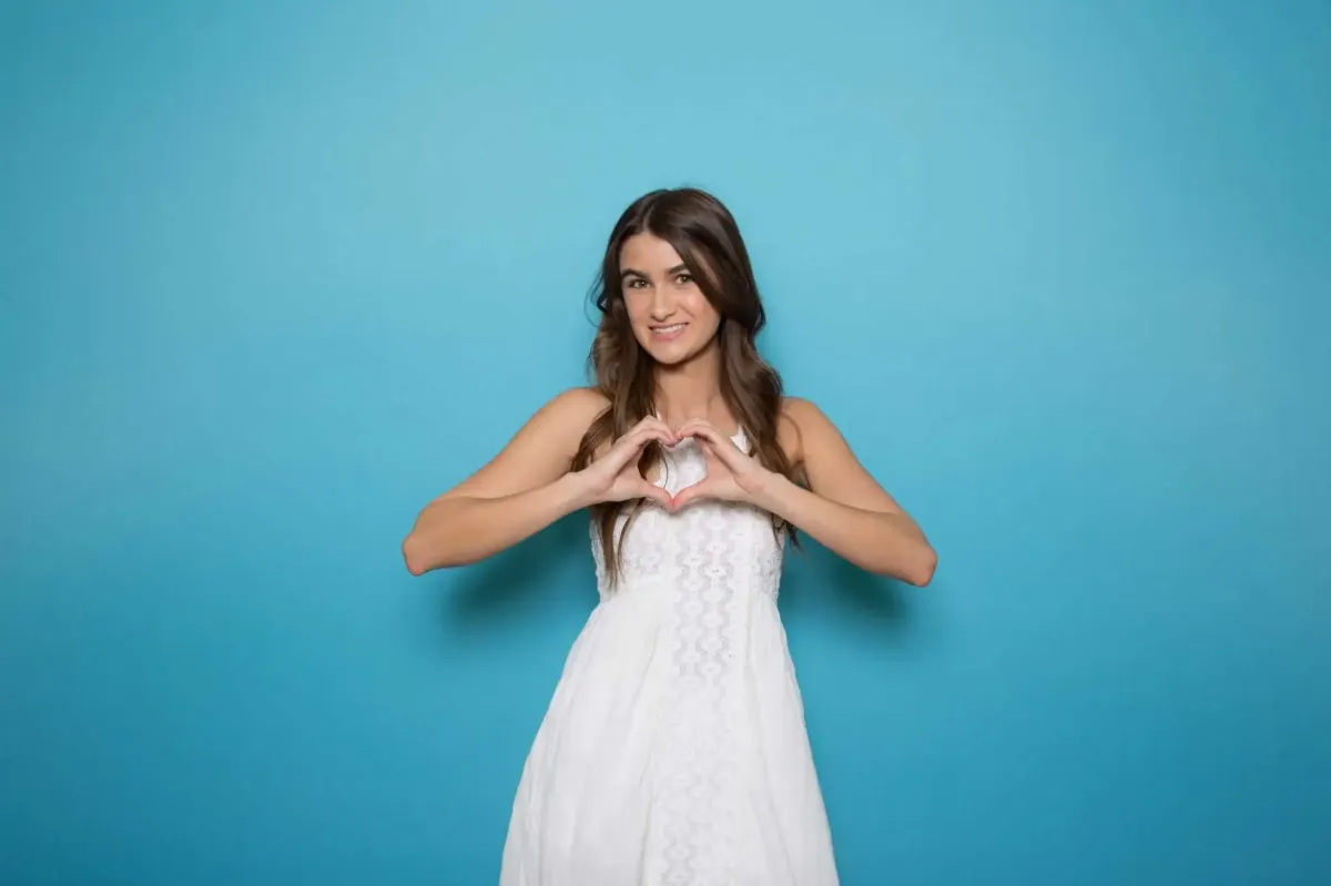 A woman in a white dress stands against a blue background, making a heart shape with her hands—perfect for your next event with Photo Booth Rental DC.