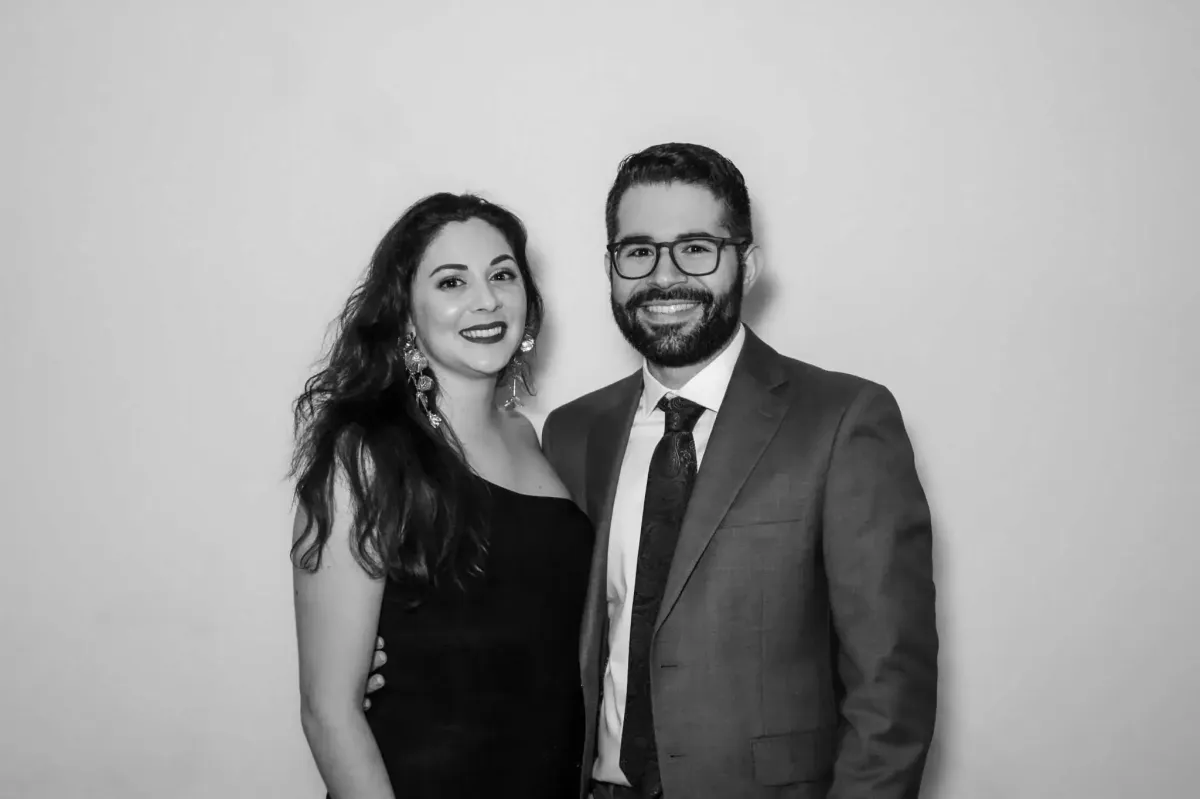 A woman in a black dress and a man in a suit and tie stand together, smiling, against a plain background, capturing the perfect moment with Photo Booth Rental DC.