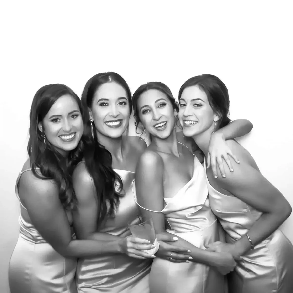 Four women in matching light-colored dresses pose closely together, smiling and embracing each other against a plain background. One holds a drink. This delightful moment captures the essence of a great Photo Booth Rental, ensuring memories are timeless.