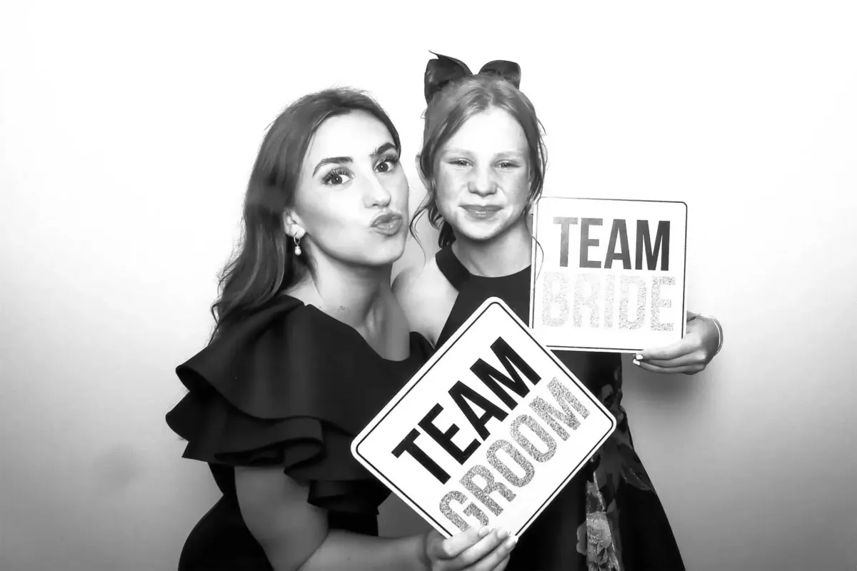 Two people posing in a Black & White Photo Booth. An adult on the left holds a "Team Groom" sign and a child on the right holds a "Team Bride" sign. Both are smiling, capturing the joy of the moment with Photo Booth Rental DC.