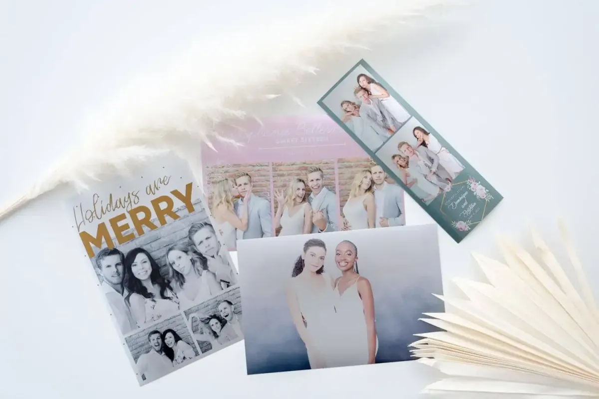 Various photo prints and holiday cards are arranged on a white surface alongside a decorative plant, an open book, and memories captured from a Photo Booth Rental.