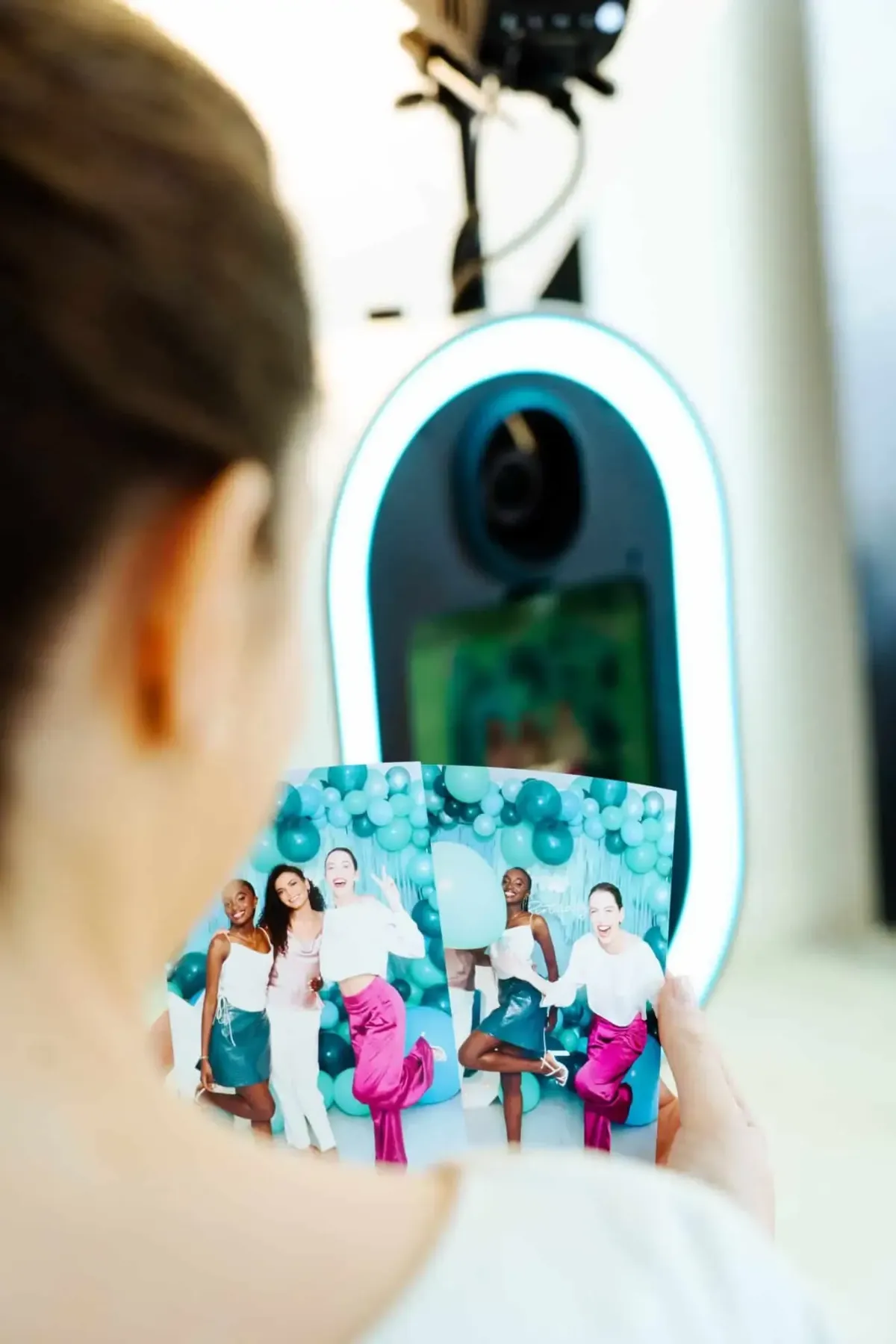 A person holds two photo booth prints from an event in DC, Maryland, or Virginia, capturing three friends posing joyfully against a vibrant blue balloon backdrop.