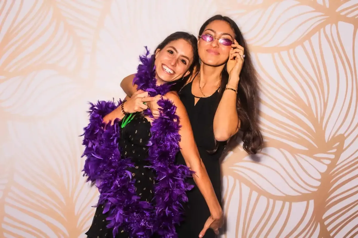 Two women smiling, one draped in a purple feather boa and the other adjusting her eyeglasses, pose playfully in front of a patterned backdrop at the corporate event's photo booth.