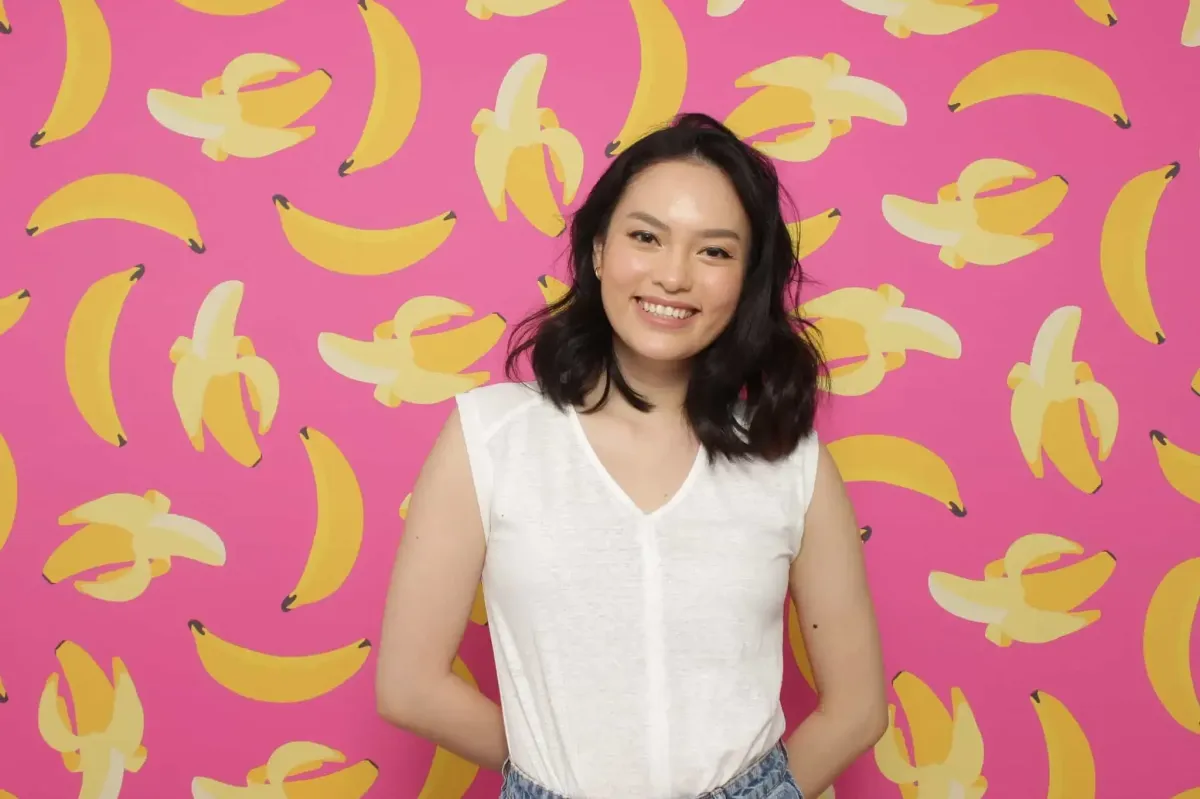 A person smiles brightly in front of a vibrant pink wall adorned with a playful banana pattern, reminiscent of a lively photo booth backdrop perfect for private events.