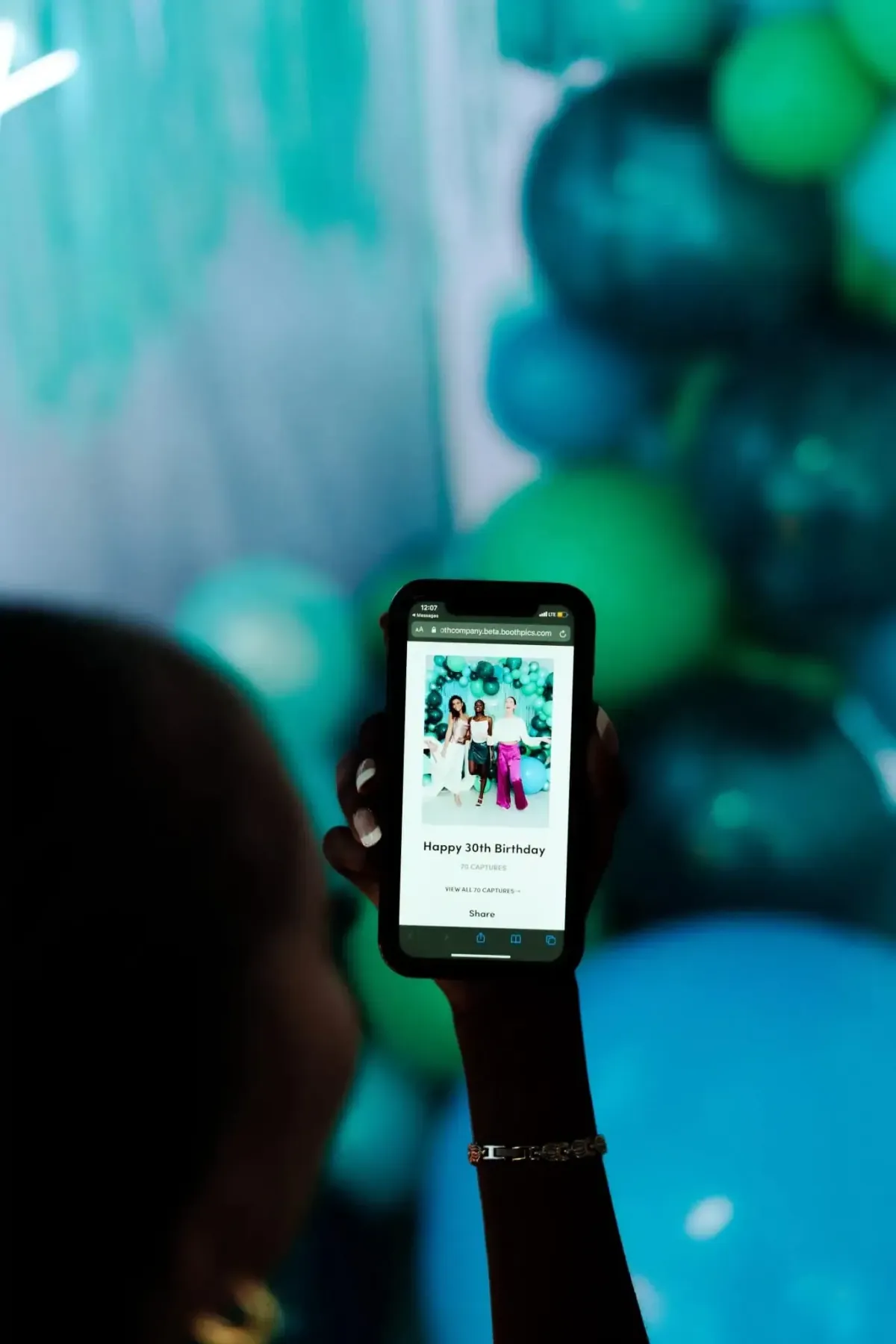 Person holding a smartphone displaying a photo with a "Happy 30th Birthday" message, in front of a backdrop with blue and green balloons from a Photo Booth Rental DC.