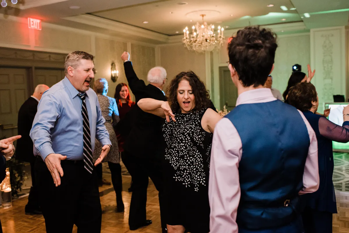 Adults dancing and laugh together at a festive indoor event with string lights, formal attire, and a Wedding DJ creating an unforgettable atmosphere in the background.