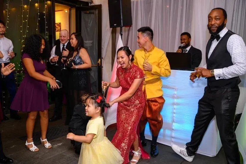 Adults and children dance and laugh together at a festive indoor event with string lights, formal attire, and a Multicultural Wedding DJ creating an unforgettable atmosphere in the background.