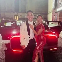 A man in a white tuxedo and a woman in a red dress stand together smiling in front of a black car with its doors open at night, ready for an unforgettable evening with their wedding DJ.