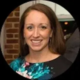 A woman with long brown hair in a floral top smiles at the camera, while a brick wall and window hint at a lively setting—perfect for capturing moments or celebrating with a Wedding DJ.