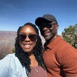 Two people wearing sunglasses pose for a selfie outdoors with trees and a canyon landscape in the background under a clear blue sky, capturing the moment after booking their favorite Wedding DJ.