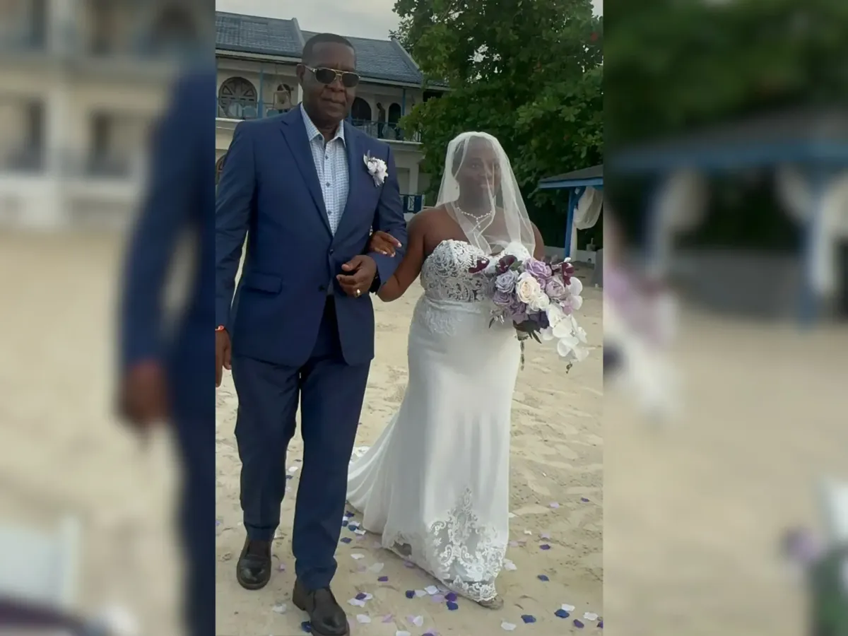 A woman in a wedding dress with a veil holds a bouquet of flowers and walks on sand with a man in a blue suit at an outdoor venue.