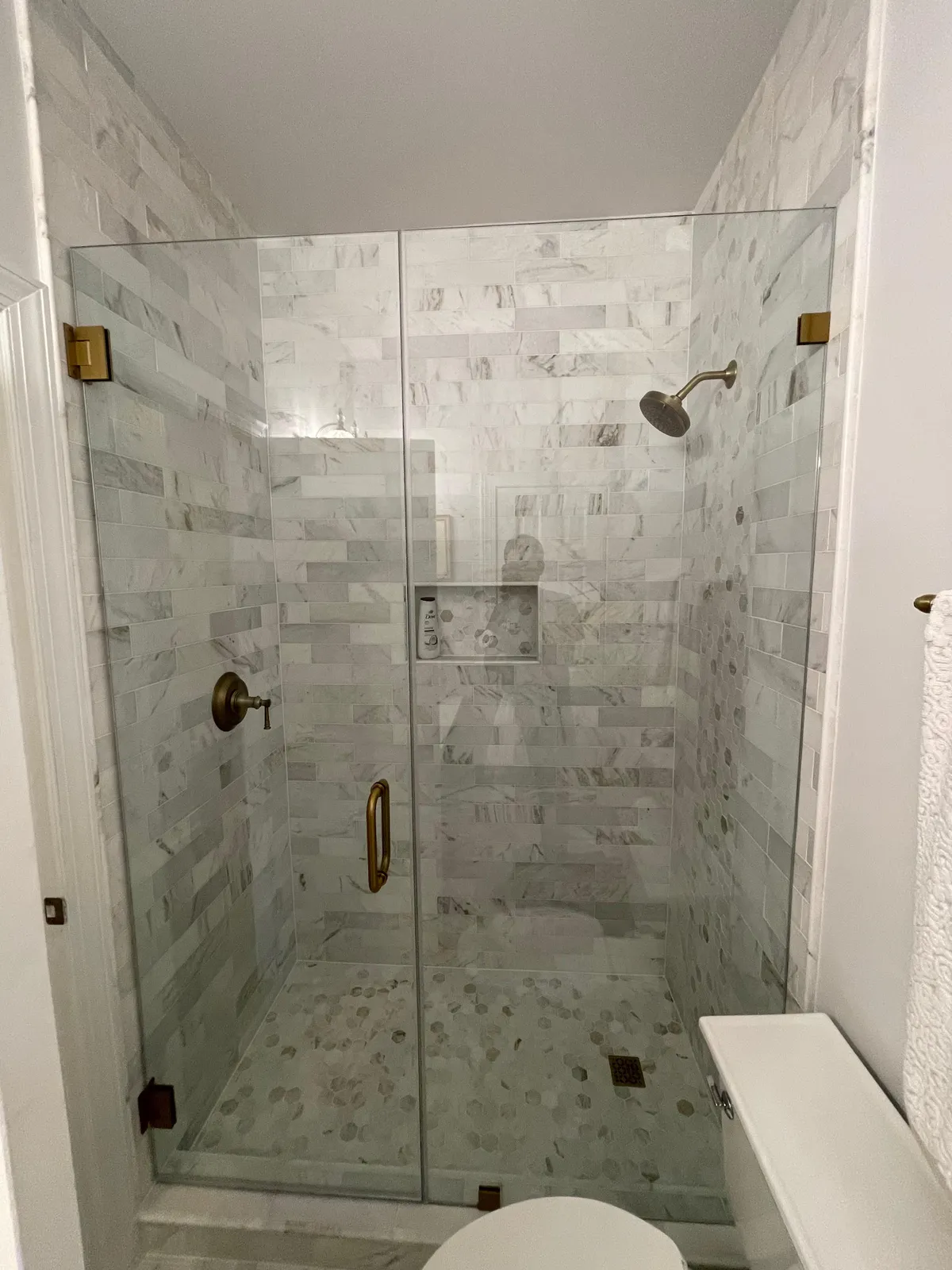 Frameless walk-in shower with marble-look tile, brass fixtures and hexagonal mosaic floor by JR Remodeling