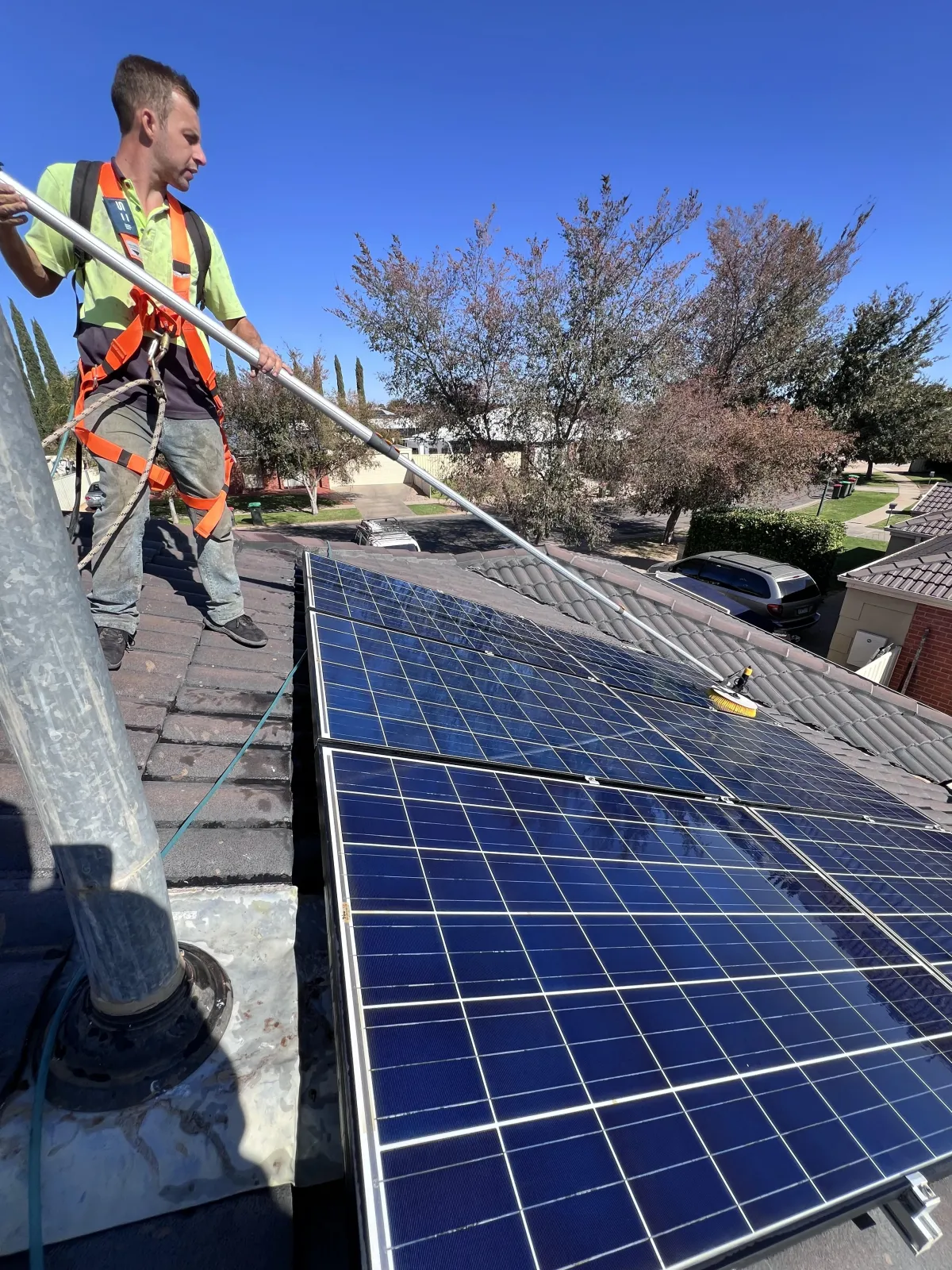 Technician in safety harness cleaning rooftop solar panels with a water-fed pole on a sunny day in Mildura.