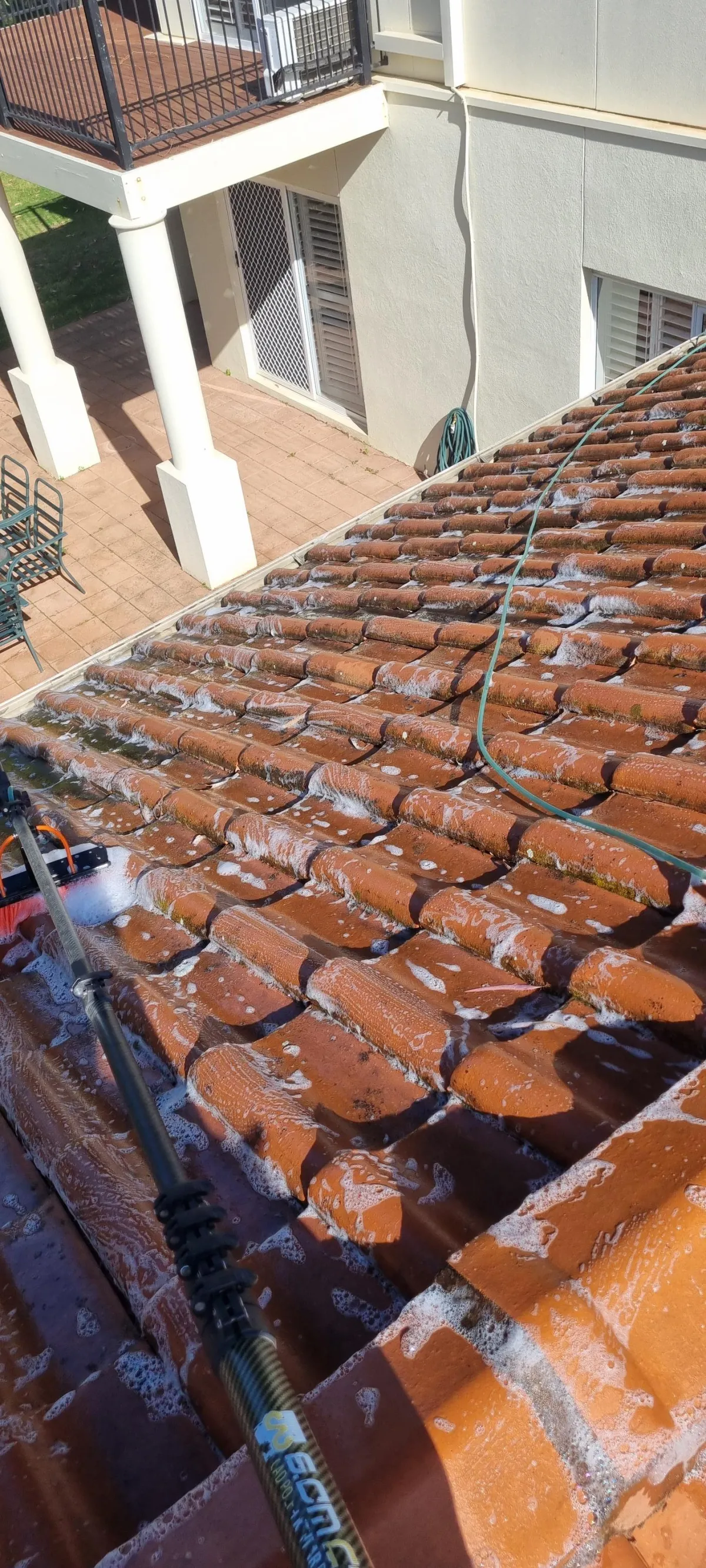 "Close-up of terracotta roof tiles being cleaned with professional equipment. The image shows soapy water flowing down the tiles during the cleaning process, with a specialized water-fed cleaning brush visible. The photo demonstrates the dramatic difference between cleaned and uncleaned sections of the tiled roof, with a patio area visible below.