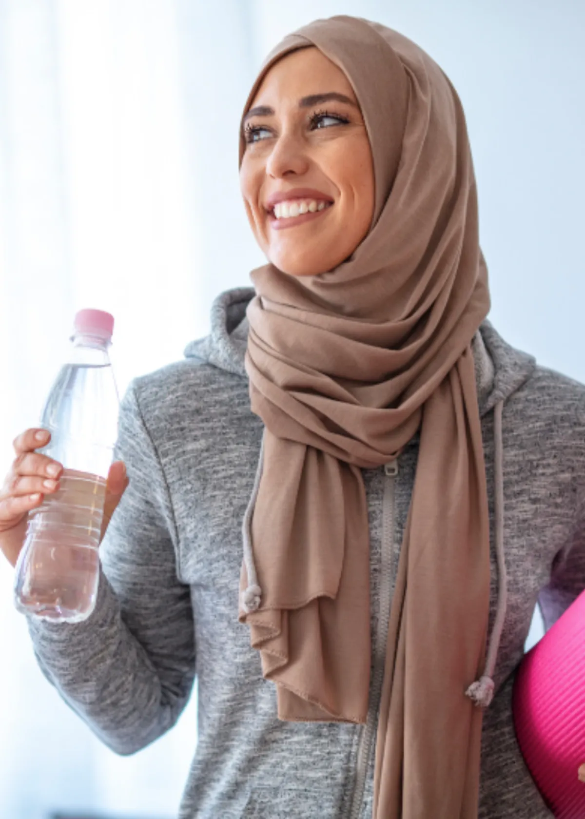 A smiling woman in a hijab holding a water bottle and a yoga mat ready for a personal trainer certification
