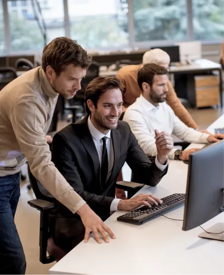 four men in an office setting wearing business attire. they are surrounding a computer screen and looking at it. one in a black suit is smiling while another is looking over his shoulder at the computer.