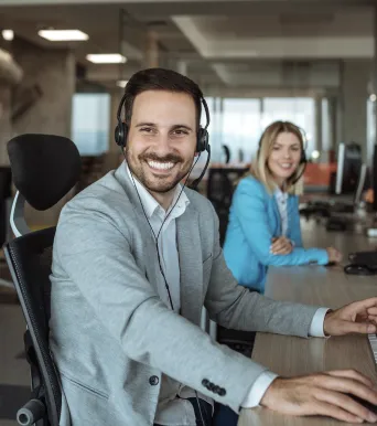 man with a suit in an office setting wearing a headset and sitting at a computer. he is looking at the camera smiling. there is a woman behind him in a blue suit with a headset on looking at the camera and smiling