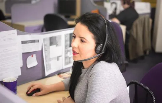 a woman in a gray shirt with black hair n an office wearing a headset looking at a computer screen with her hand on a mouse