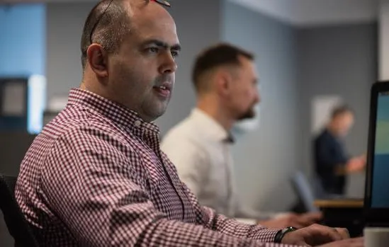 A man in a maroon button up with glasses on his forehead sitting in an office and looking at a computer screen. behind him is a man in a white button up looking at a computer screen. 