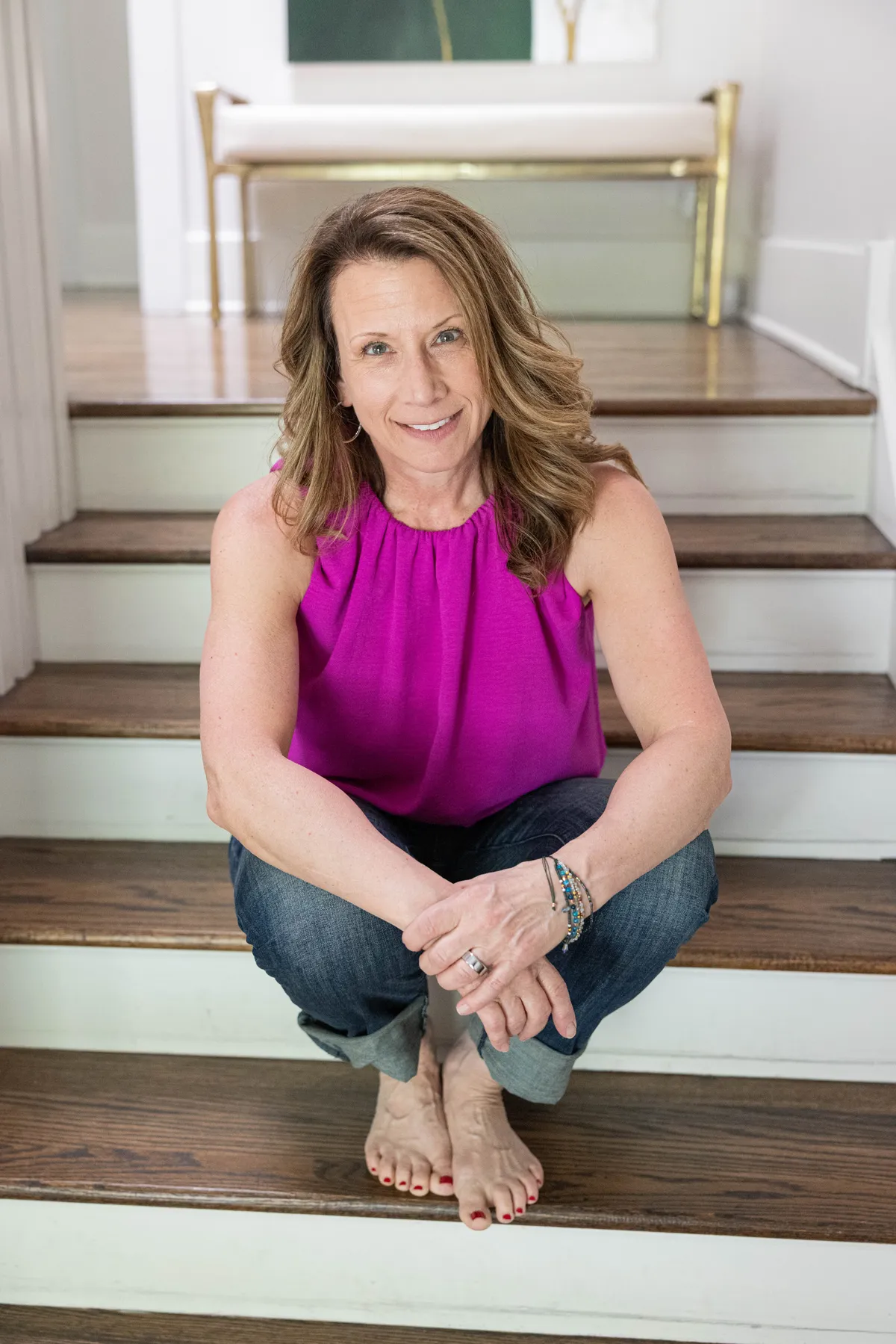 Certified health coach and movement specialist sitting on wooden stairs in a bright home, wearing a purple sleeveless top and jeans, representing strength, mobility, and wellness