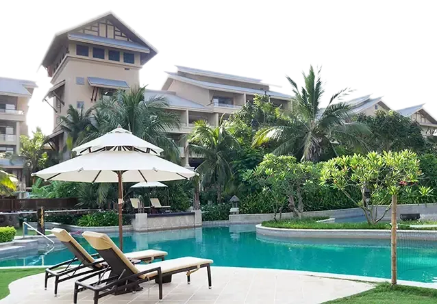 A resort-style swimming pool area with two lounge chairs under an umbrella, surrounded by lush greenery and multi-story buildings in the background, meticulously maintained by Columbia Master Services.