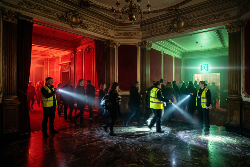 A Victorian theatre foyer T-junction during a low-light evacuation. Left corridor in red, right corridor in green, two stewards with torch beams cutting through atmospheric haze.