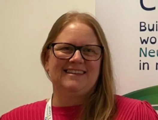 Lucy Smith, a smiling woman wearing glasses and a bright pink top, and founder and director of Inclusive Change, standing in front of an Inclusive banner.