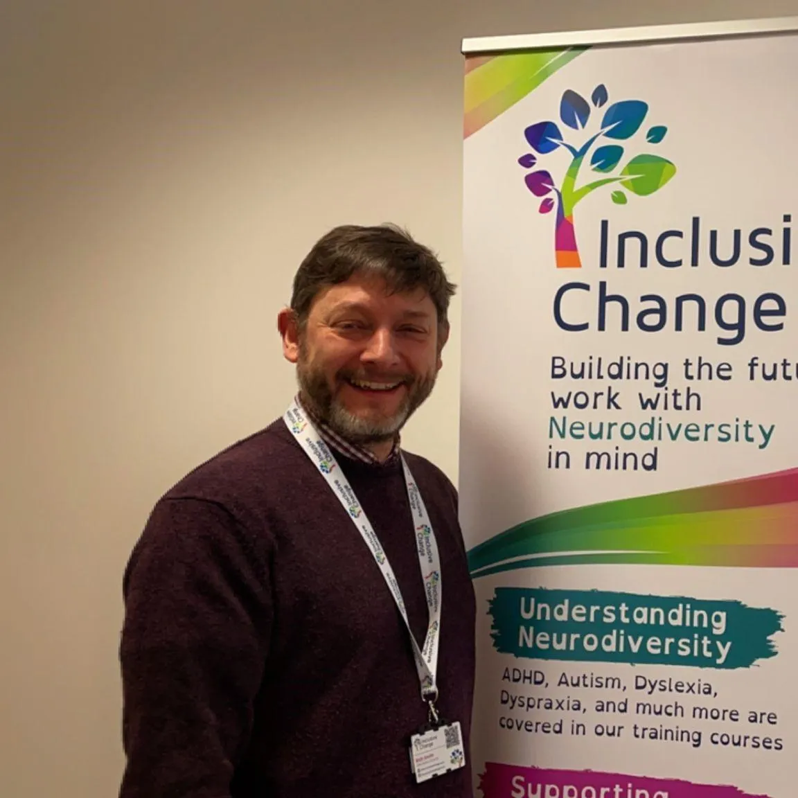 Richard Smith, a smiling man with short dark hair and a bear, wearing a maroon jumper over a chequered shirt and a lanyard, and operations director of Inclusive Change, standing in front of an Inclusive Change banner.