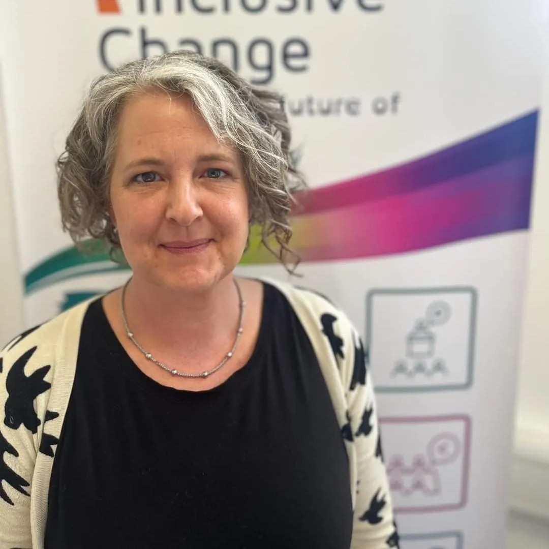 Emily Chittell, a woman with short, curly grey hair wearing a black top and a cream jumper with a black bird pattern, project manager of Inclusive Change, standing in front of an Inclusive Change banner.